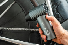 Eine Person benutzt das „Optimize Akku Luftgebläse“ der Optimize GmbH – ein kompaktes, graues Gerät mit der Aufschrift „OPTIMIZE“ – in der Nähe einer Fahrradkette, vermutlich, um Schmutz effizient wegzublasen und das Fahrrad zu trocknen.