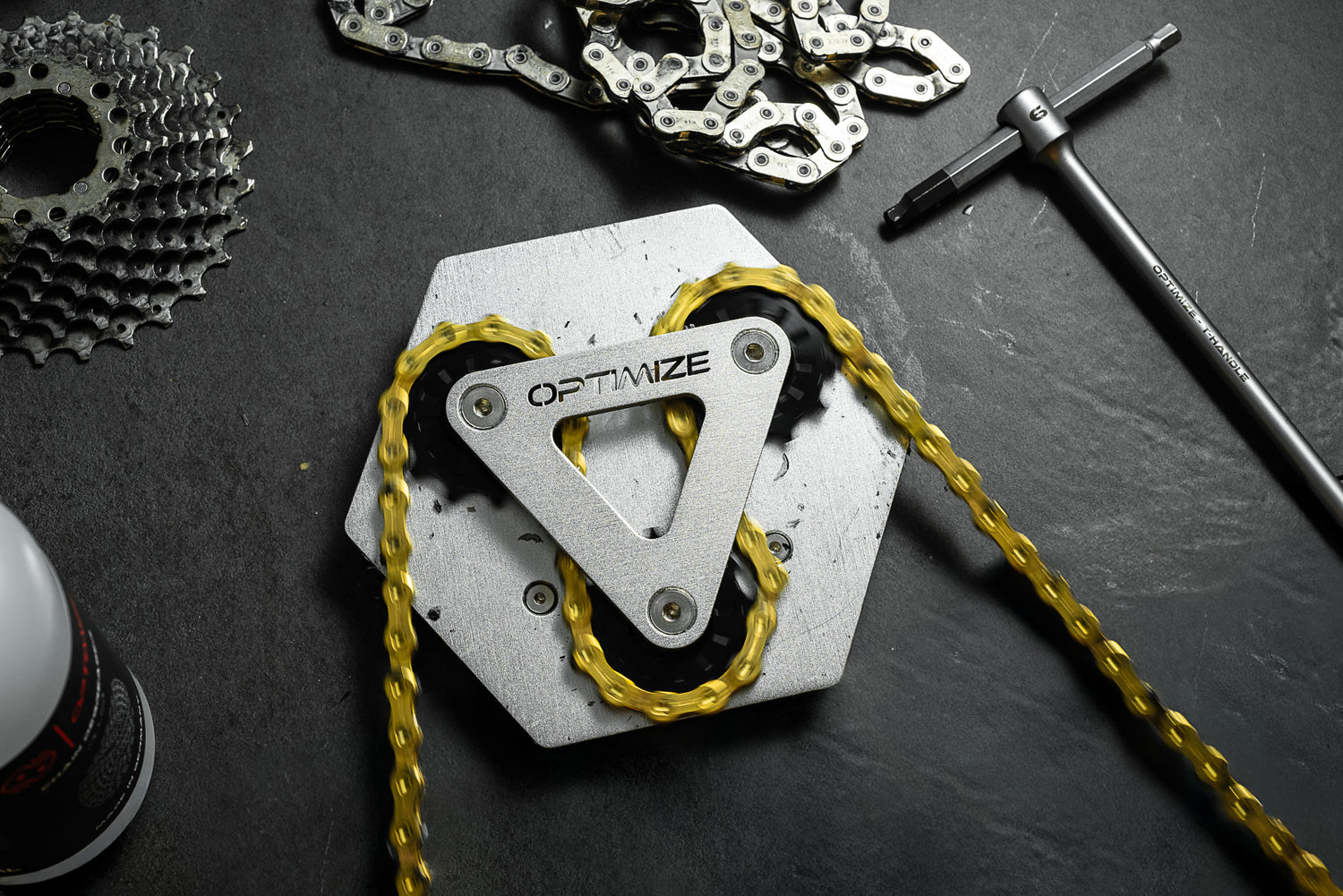 Eine Nahaufnahme des silbernen Kettenreinigungswerkzeugs „Kettenbrecher mechanisch“ der Optimize GmbH, durch das eine goldene Kette geschlungen ist, umgeben von einem Zahnradsatz und weiteren Werkzeugen, präsentiert auf einem dunklen Untergrund.