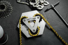 Eine Nahaufnahme des silbernen Kettenreinigungswerkzeugs „Kettenbrecher mechanisch“ der Optimize GmbH, durch das eine goldene Kette geschlungen ist, umgeben von einem Zahnradsatz und weiteren Werkzeugen, präsentiert auf einem dunklen Untergrund.