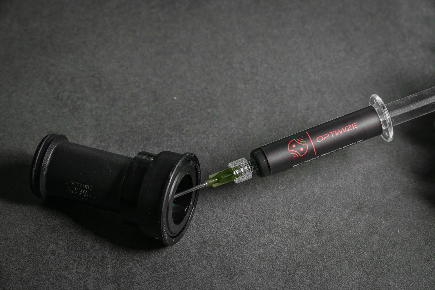 Eine Spritze mit der Aufschrift Optimize injiziert grünen Kettenwachs in ein schwarzes zylindrisches Fahrradteil, möglicherweise ein Tretlager, vor einem dunkelgrauen Hintergrund.