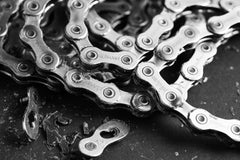 Eine Nahaufnahme in Schwarz-Weiß zeigt die Connex 12SR WAX - gewachste 12-fach-Kette für SRAM Flattop der Optimize GmbH auf einer dunklen Fläche, auf einem Glied ist "GERMANY" eingraviert, daneben liegen kleine Metallfragmente.