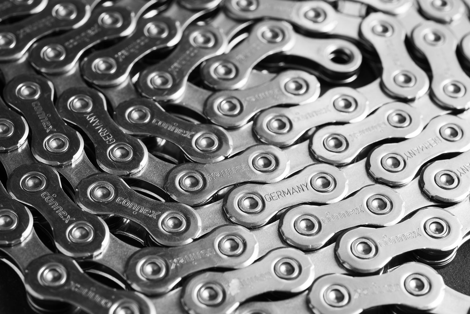 Nahaufnahme von ineinander greifenden Fahrradketten aus Metall, darunter die Optimize GmbH Connex 12SR WAX - eine gewachste 12-fach-Kette für SRAM Flattop, mit sichtbaren Bolzen und eingraviertem Text, der detaillierte, polierte Glieder in einem sich wiederholenden Muster hervorhebt.