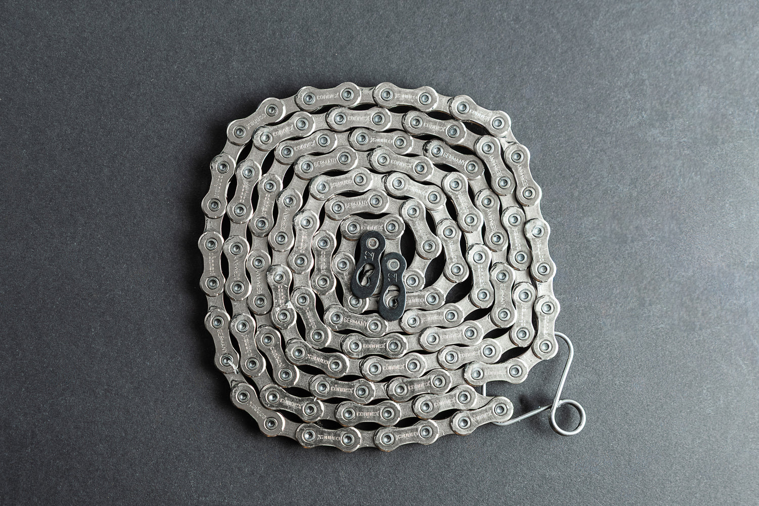 Eine gewickelte Connex 12SR WAX - gewachste 12-fach-Kette für SRAM Flattop von Optimize GmbH sitzt in einer engen Spirale auf einer dunkelgrauen Fläche, Quicklink nahe der Mitte und das Kettenende leicht nach außen verlängert.